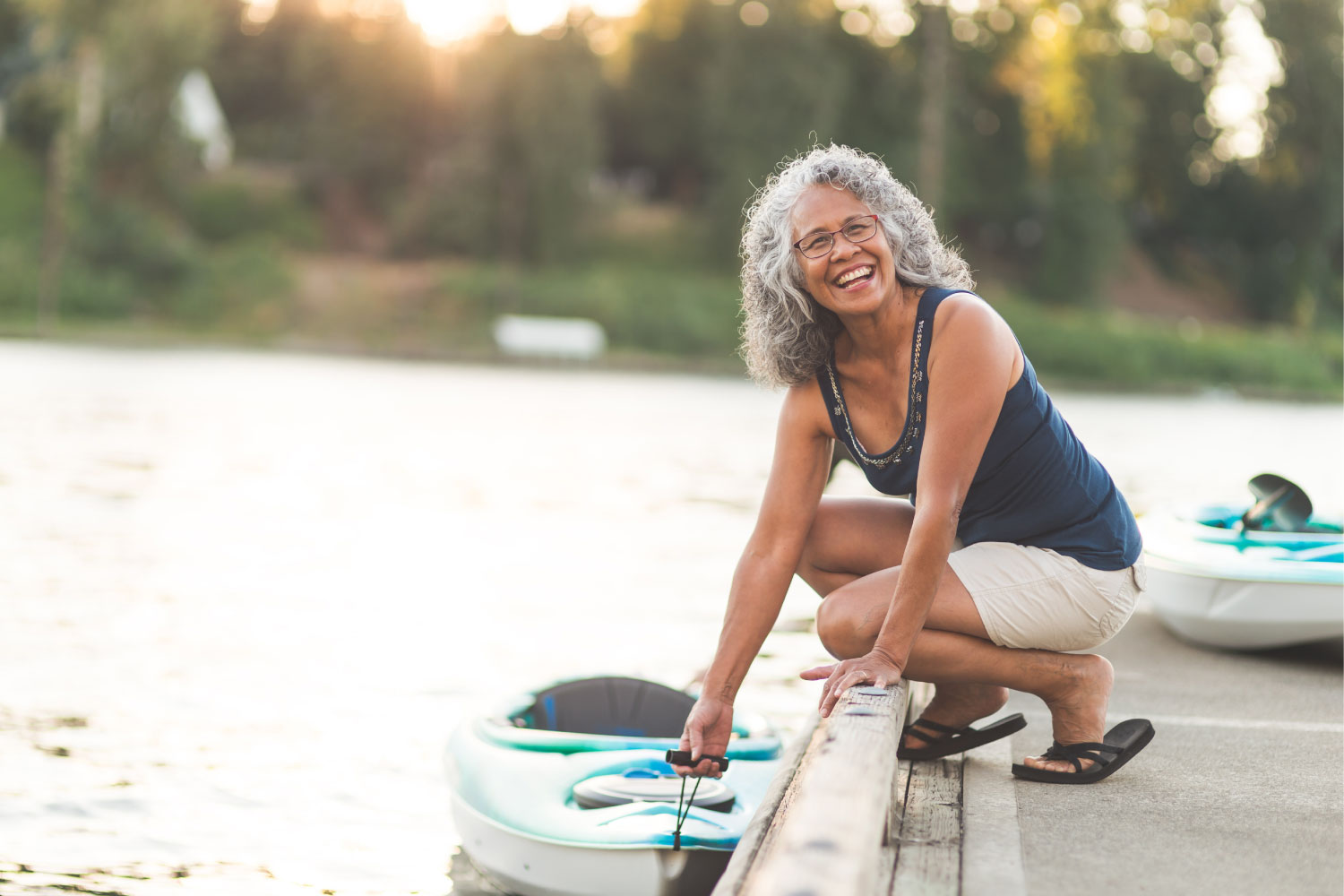 Woman with dentures smiles while holding her kayak on a lake after receiving care at Rockingham Prosthodontics