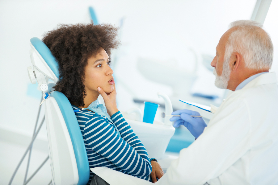 Black woman with curly hair touches her chin while sitting in a dental chair and consults with her middle-age male dentist