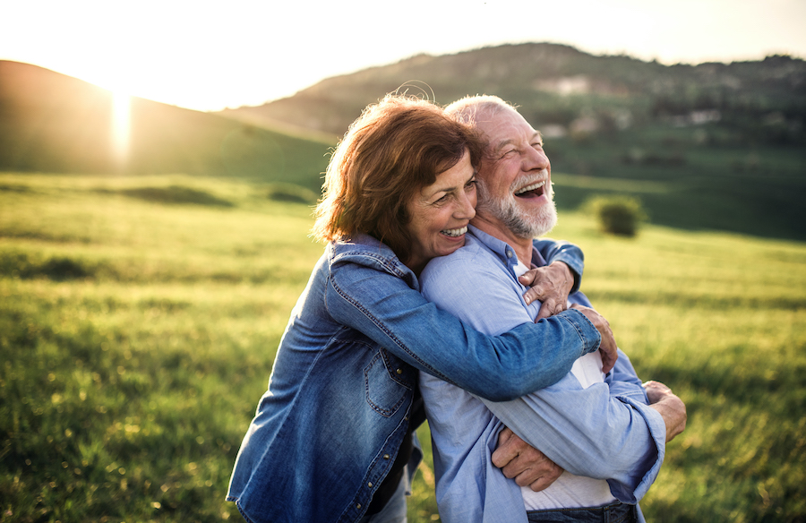 A middle-aged woman and man smile as they embrace in a grassy meadow as the sun sets