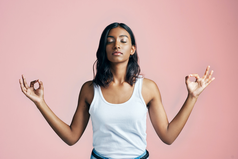 Brown woman in a white tanktop stands relaxed in a zen pose against a pink background