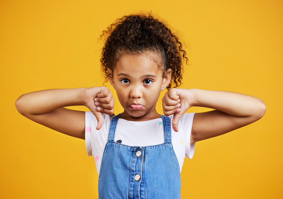 Adorable young Black girl in a denim dress frowns and has 2 thumbs down against a yellow background