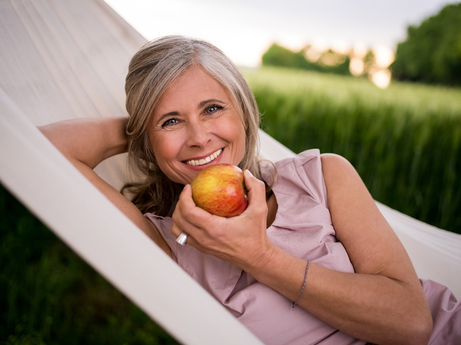 Healthy mature woman relaxing in a hammock in a park and eating a fresh apple with a healthy smile
